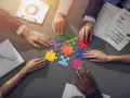 Des mains assemblent des pièces de puzzle colorées, symbolisant la collaboration en équipe. Des graphiques et un bloc-notes complètent la scène.