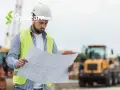 Un chef de projet AMOA, casque blanc et gilet fluo, examine des plans sur un chantier.