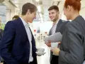 Trois personnes en costume discutent avec aisance relationnelle autour d'un café. L'un tient des dossiers.