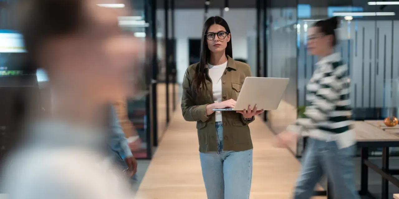 Une program manager concentrée, portable à la main, traverse un couloir d'entreprise moderne.