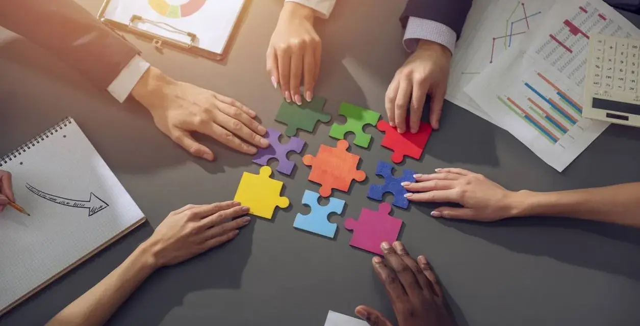 Des mains assemblent des pièces de puzzle colorées, symbolisant la collaboration en équipe. Des graphiques et un bloc-notes complètent la scène.