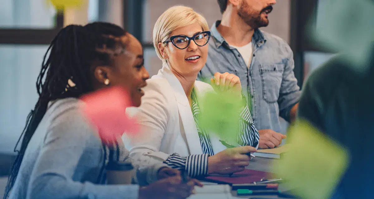 Une équipe discute autour d'une table, la chef de projet souriante au centre. Des post-its colorés flottent, symbolisant la créativité et la collaboration.