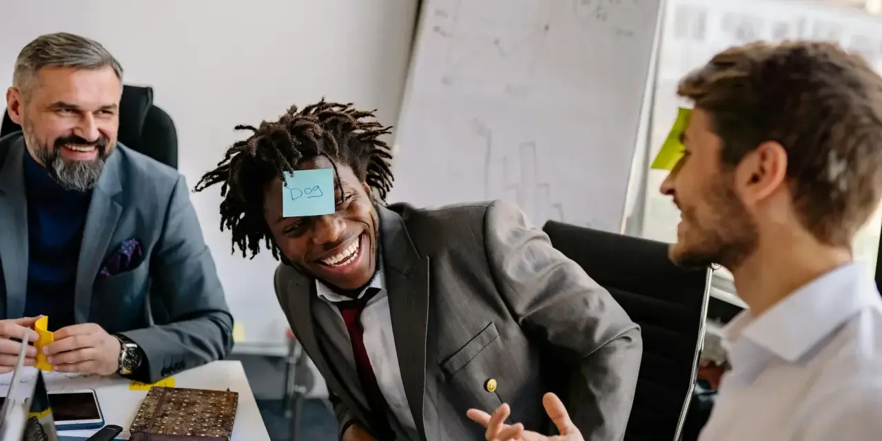 Un homme rit avec une note "Dog" sur le front, participant à un jeu de communication d'équipe.