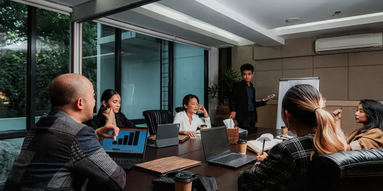 Une équipe discute de stratégies de management et communication autour d'une table de réunion. Un homme présente des graphiques sur un écran.
