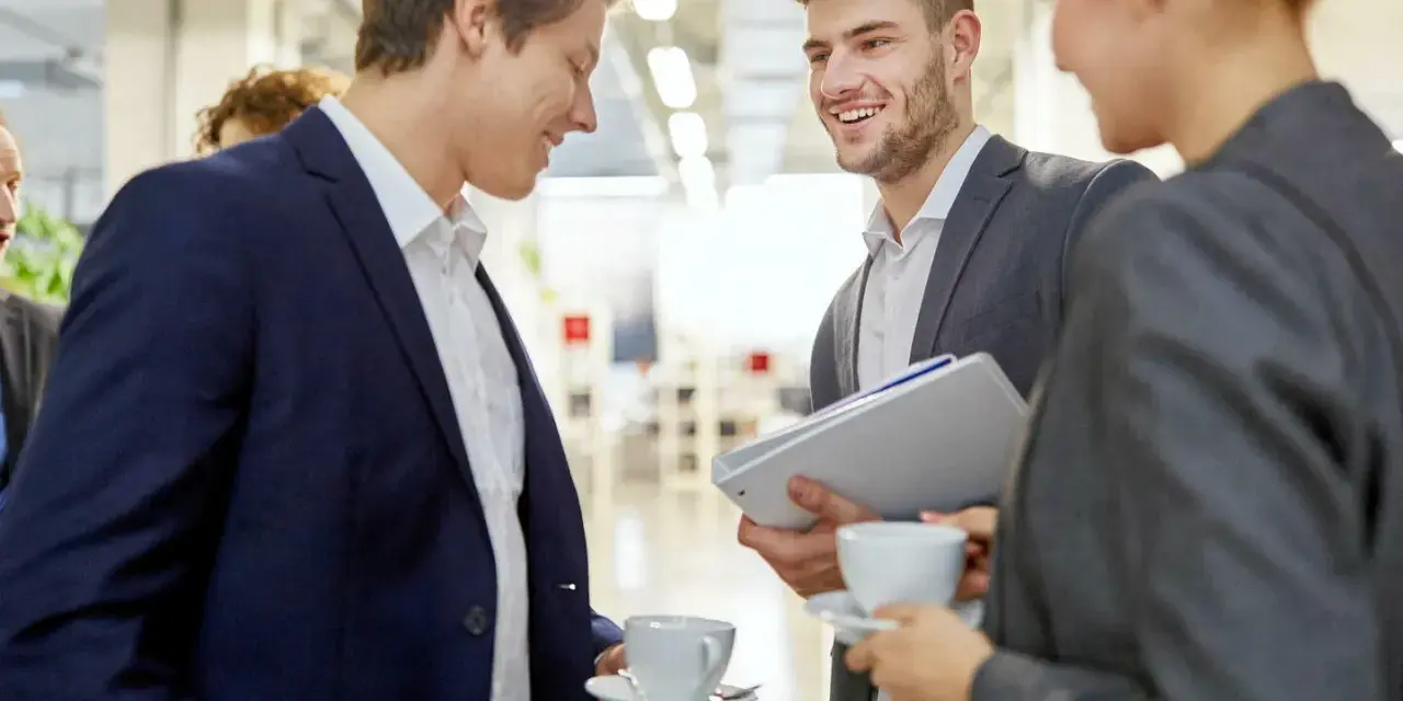 Trois personnes en costume discutent avec aisance relationnelle autour d'un café. L'un tient des dossiers.