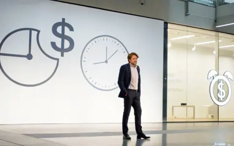 Un homme en costume présente une conférence pecha kucha. Des horloges et un symbole dollar sur l'écran suggèrent le thème du temps et de l'argent.