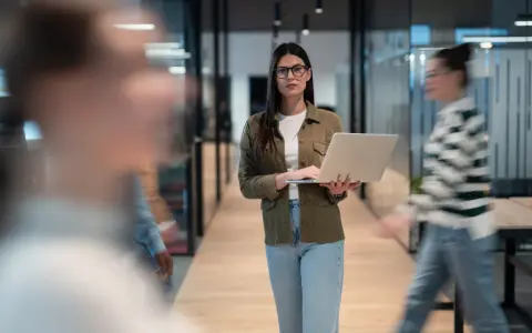 Une program manager concentrée, portable à la main, traverse un couloir d'entreprise moderne.
