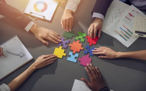 Des mains assemblent des pièces de puzzle colorées, symbolisant la collaboration en équipe. Des graphiques et un bloc-notes complètent la scène.