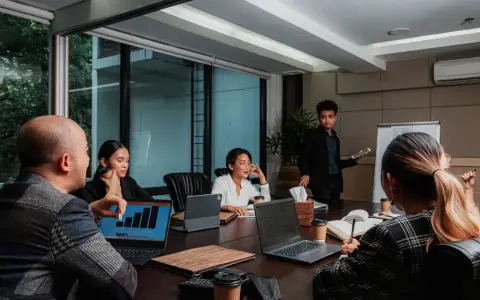 Une équipe discute de stratégies de management et communication autour d'une table de réunion. Un homme présente des graphiques sur un écran.