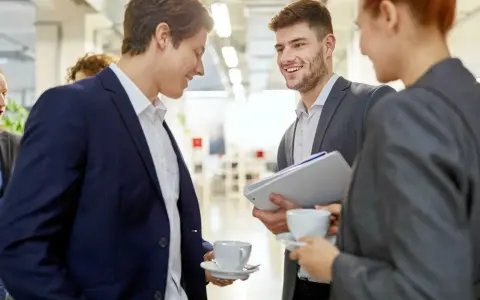 Trois personnes en costume discutent avec aisance relationnelle autour d'un café. L'un tient des dossiers.