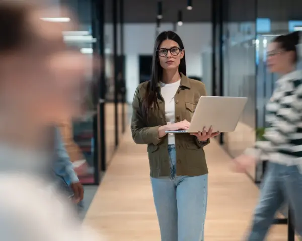 Une program manager concentrée, portable à la main, traverse un couloir d'entreprise moderne.