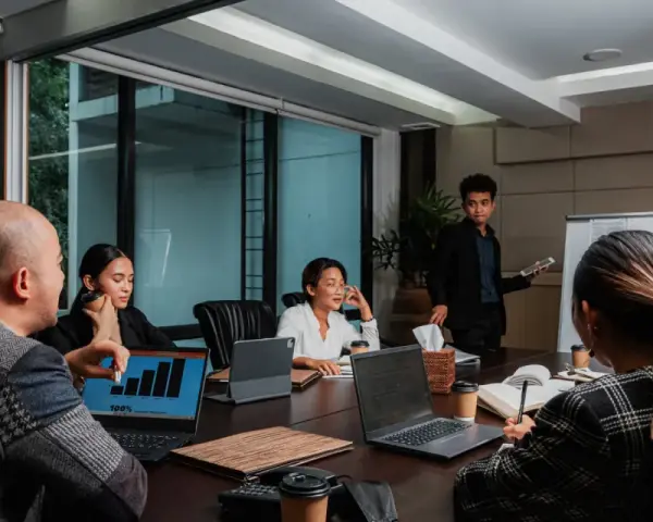 Une équipe discute de stratégies de management et communication autour d'une table de réunion. Un homme présente des graphiques sur un écran.