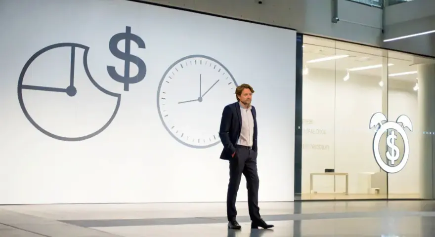 Un homme en costume présente une conférence pecha kucha. Des horloges et un symbole dollar sur l'écran suggèrent le thème du temps et de l'argent.