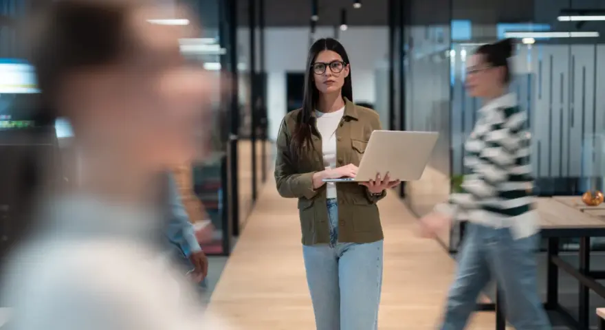 Une program manager concentrée, portable à la main, traverse un couloir d'entreprise moderne.