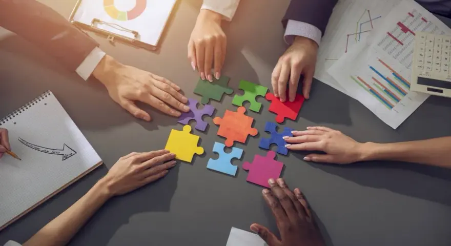 Des mains assemblent des pièces de puzzle colorées, symbolisant la collaboration en équipe. Des graphiques et un bloc-notes complètent la scène.
