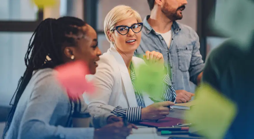 Une équipe discute autour d'une table, la chef de projet souriante au centre. Des post-its colorés flottent, symbolisant la créativité et la collaboration.