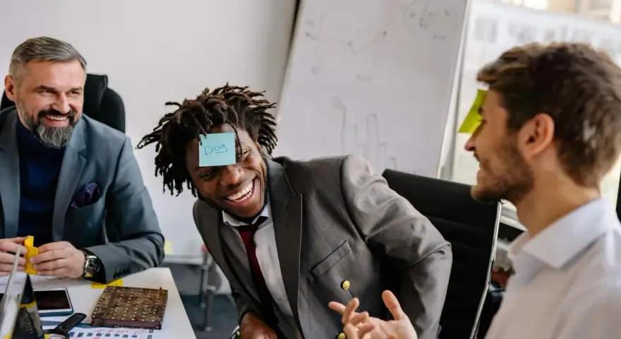 Un homme rit avec une note "Dog" sur le front, participant à un jeu de communication d'équipe.