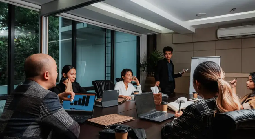 Une équipe discute de stratégies de management et communication autour d'une table de réunion. Un homme présente des graphiques sur un écran.