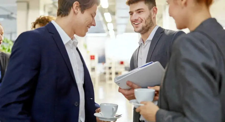 Trois personnes en costume discutent avec aisance relationnelle autour d'un café. L'un tient des dossiers.