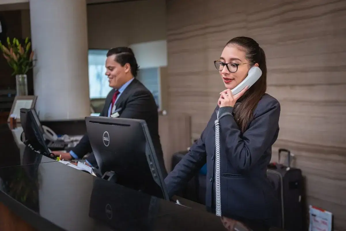 Une r&eacute;ceptionniste au t&eacute;l&eacute;phone g&egrave;re l'accueil t&eacute;l&eacute;phonique d'une entreprise. Un coll&egrave;gue travaille sur un ordinateur &agrave; c&ocirc;t&eacute;.