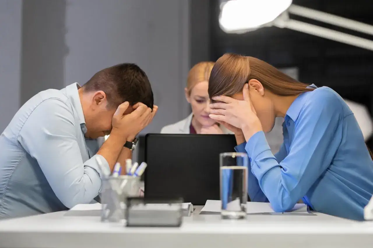 Trois employ&eacute;s accabl&eacute;s, mains sur le visage, face &agrave; un &eacute;cran d'ordinateur. L'ambiance sugg&egrave;re un mauvais manager et une surcharge de travail.