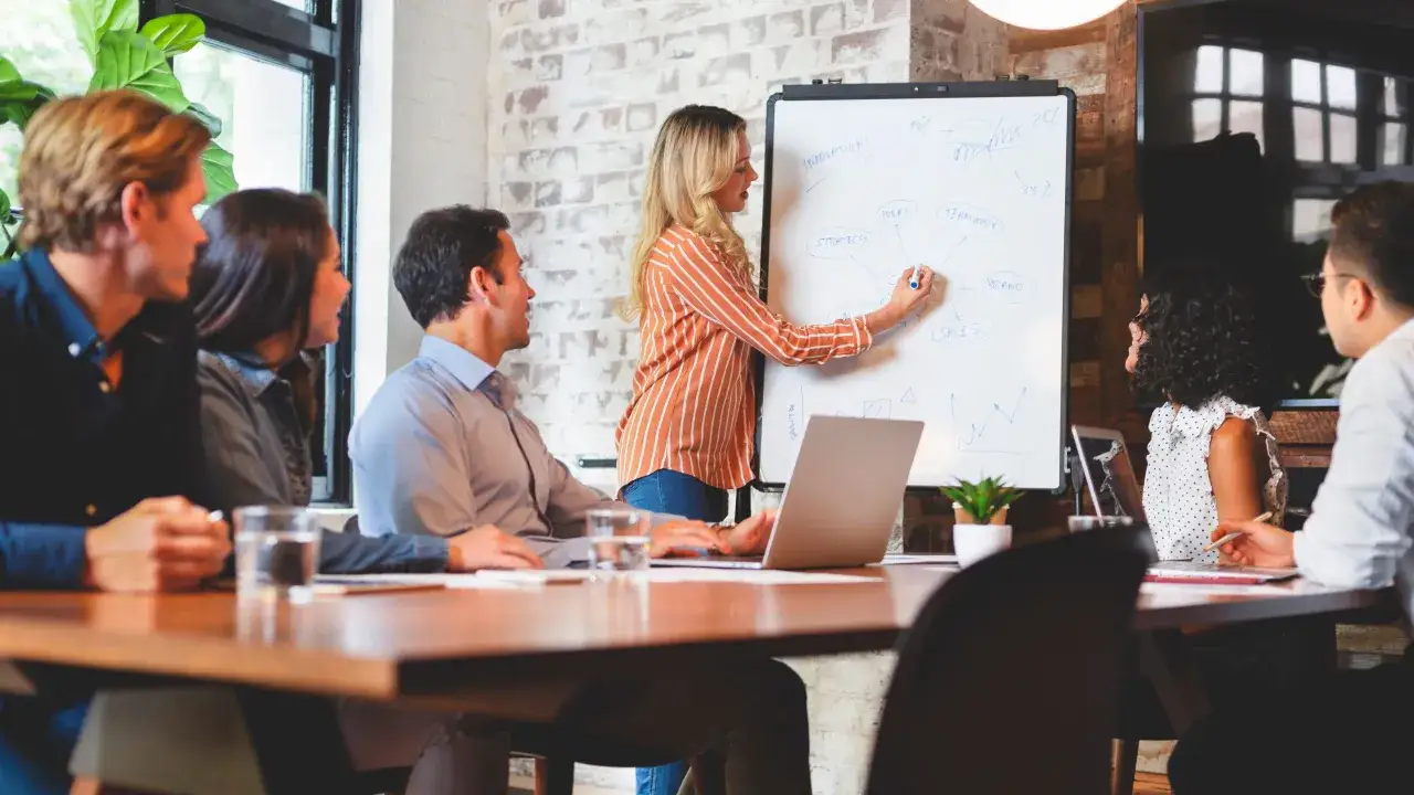 Une équipe discute stratégie et innovation. La formatrice, au tableau blanc, illustre avec esprit de synthèse les idées clés : stratégie, teamwork, ventes, marque.