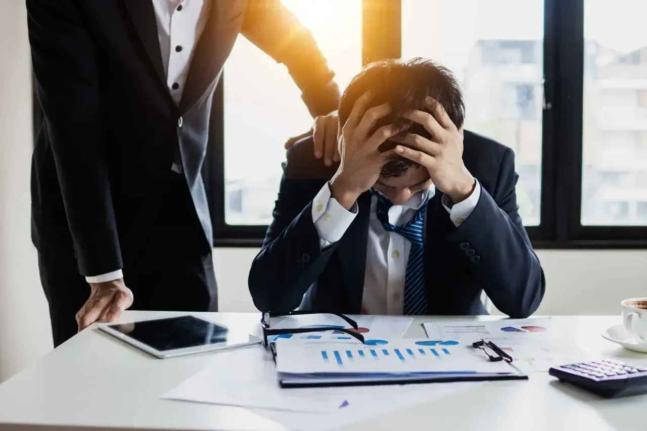 Un homme d'affaires désespéré, accablé par le stress, reçoit une tape sur l'épaule. L'ambiance suggère un management toxique.