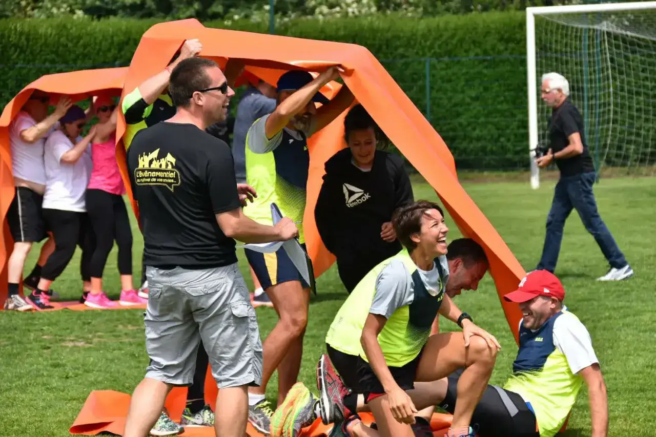 Des participants hilares lors d'un jeu de team building jeux olympiques sous un grand tissu orange.