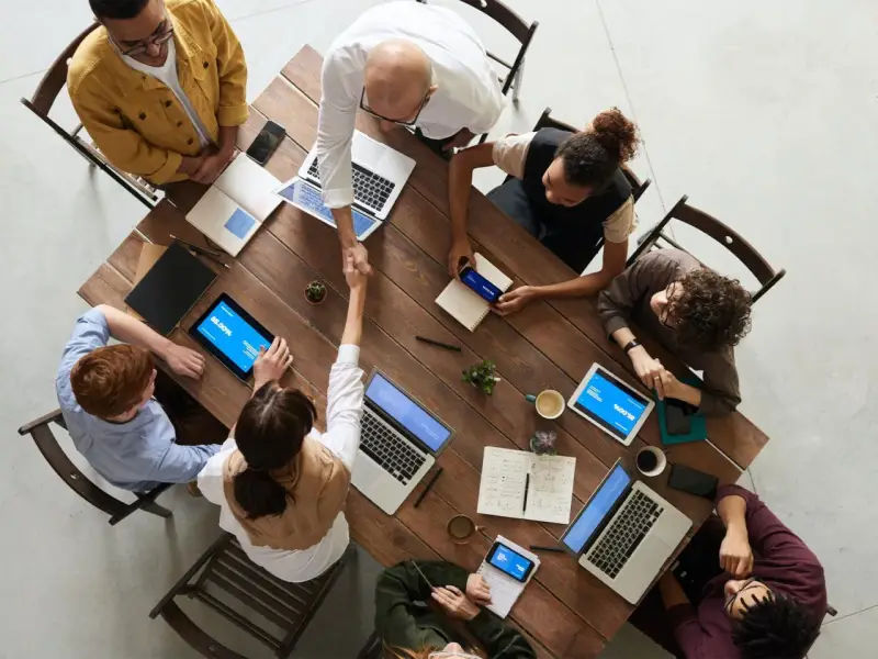Une équipe diversifiée collabore autour d'une table en bois, renforçant la communication et la cohésion d'équipe par des poignées de main et des discussions.
